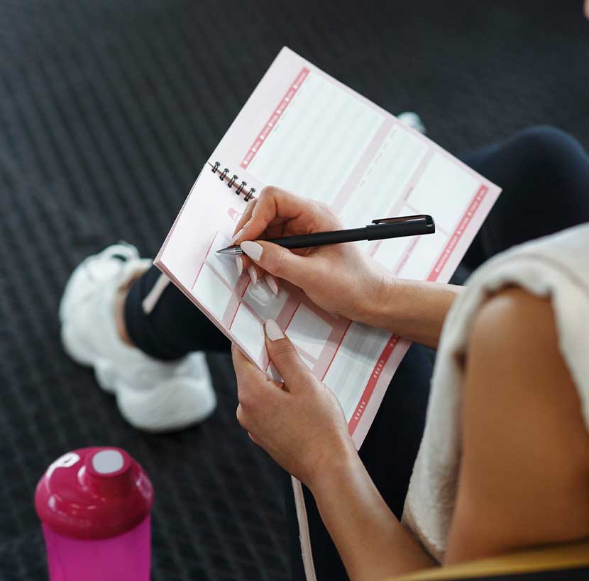A imagem mostra uma mulher a escrever num caderno o seu novo plano de treinos no ginásio.