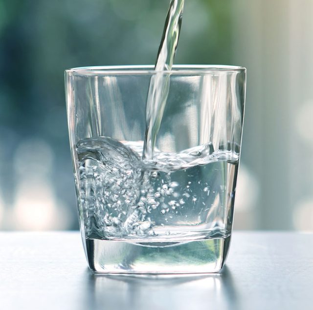 Se llena un vaso transparente con agua, creando pequeñas burbujas y ondas. El fondo está borroso, dando la sensación de un ambiente luminoso y bien iluminado. La atención se centra en el vaso y el agua que se vierte en él, lo que te anima a beber más agua para una mejor hidratación.