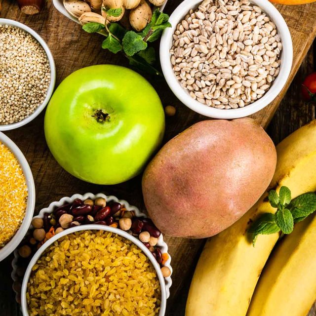 Sobre una superficie de madera se muestra una variedad de alimentos saludables perfectos para cargar carbohidratos, que incluyen una manzana verde, una papa, dos plátanos y varios tazones pequeños que contienen quinua, cebada, bulgur, frijoles y una variedad de nueces. También está presente una pequeña ramita de menta verde.