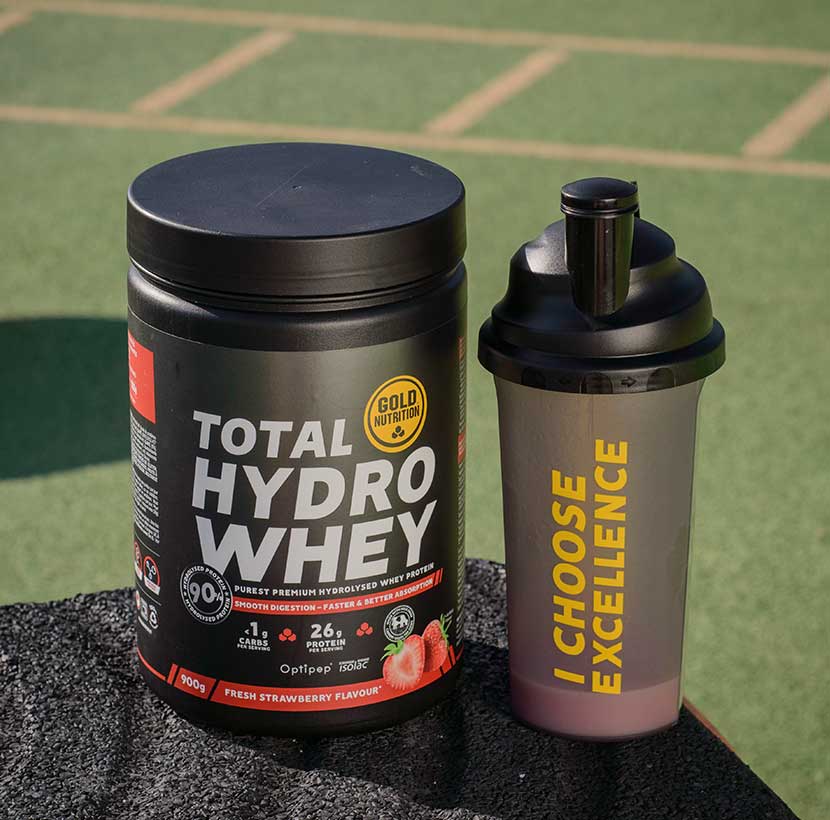 Suplemento de proteína de suero Total Hydro Whey de la marca Gold Nutrition, sabor fresa fresca, al lado de un shaker negro y gris con las palabras 'I Choose Excellence' en destaque. La imagen fue tomada al aire libre en un campo de entrenamiento | Total Hydro Whey