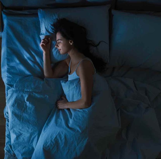 Una mujer de cabello oscuro duerme plácidamente de lado en una cama con sábanas y fundas de almohada de color azul claro, parcialmente cubierta por una manta de color azul claro. La habitación está poco iluminada, creando un ambiente sereno y tranquilo.