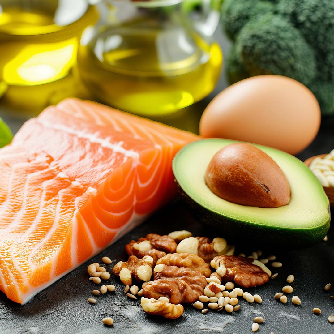 Una selección de alimentos saludables ricos en grasas buenas, incluyendo un filete de salmón crudo, medio aguacate, nueces, semillas, un huevo y botellas de aceite de oliva al fondo. La imagen muestra ingredientes frescos dispuestos sobre una superficie oscura