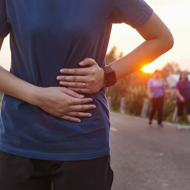 Una persona que viste una camisa azul y un reloj inteligente se sostiene el estómago con ambas manos y parece sentirse incómodo. Están al aire libre en un parque durante el atardecer, con otras dos personas borrosas en el fondo.