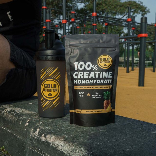 Una botella mezcladora Gold Nutrition y un paquete de monohidrato de creatina 100% Gold Nutrition se colocan sobre una superficie de concreto. Una persona sentada cerca y al fondo se ve un equipo de fitness al aire libre. El paquete de creatina tiene la etiqueta "Sabor Piña" | Creatine Monohydrate