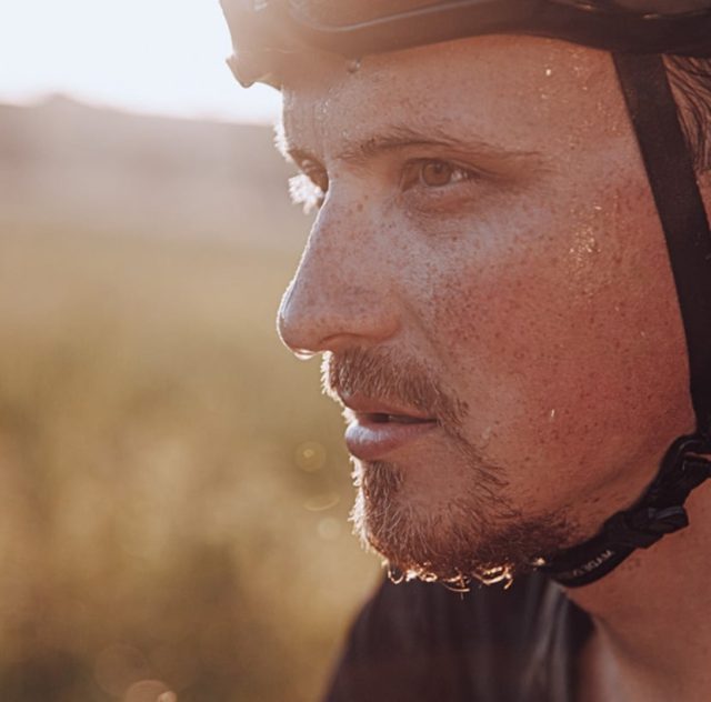 Primer plano de un hombre que lleva casco de bicicleta, con sudor visible en el rostro y una expresión solemne, que sugiere un esfuerzo físico. El fondo está borroso y la cálida luz del sol crea un suave brillo dorado.