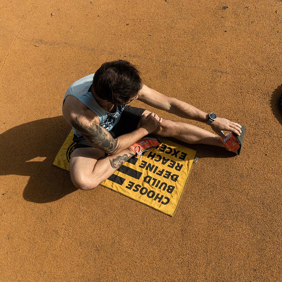 En la imagen se ve a un hombre sentado sobre una superficie naranja mientras estira una de sus piernas. Lleva una camiseta sin mangas, shorts y zapatillas deportivas naranjas. Tiene tatuajes en el brazo derecho y usa un reloj en la muñeca izquierda. Está sentado sobre una toalla amarilla con letras negras que dicen "CHOOSE BUILD DEFINE REACH EXCE". La imagen parece capturar un momento de ejercicio o estiramiento.