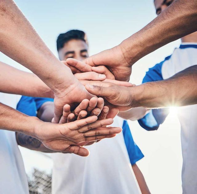 Grupo de personas, posiblemente miembros de un equipo deportivo, colocando sus manos una sobre la otra en un gesto de unidad o trabajo en equipo. Están usando camisetas deportivas, y la luz del sol brilla detrás de ellos, destacando el sentimiento de colaboración y camaradería.