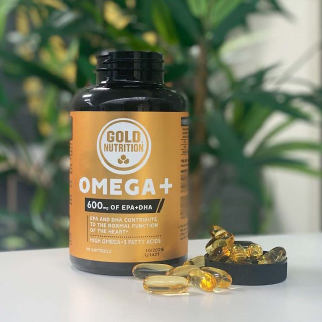 Suplemento de Omega+ de la marca Gold Nutrition, con 600 mg de EPA y DHA. Cápsulas blandas que contribuyen al funcionamiento normal del corazón, ricas en ácidos grasos Omega-3. Frasco de 90 cápsulas junto a algunas cápsulas sueltas sobre una superficie blanca, con un fondo desenfocado de plantas verdes-omega-3