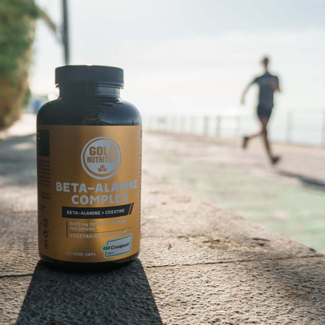 Un frasco de suplemento nutricional etiquetado como Beta-Alanina Complex de Gold Nutrition está en primer plano sobre una superficie de asfalto, mientras una persona corre en el fondo en un camino al aire libre junto al mar | Beta-Alanine Complex
