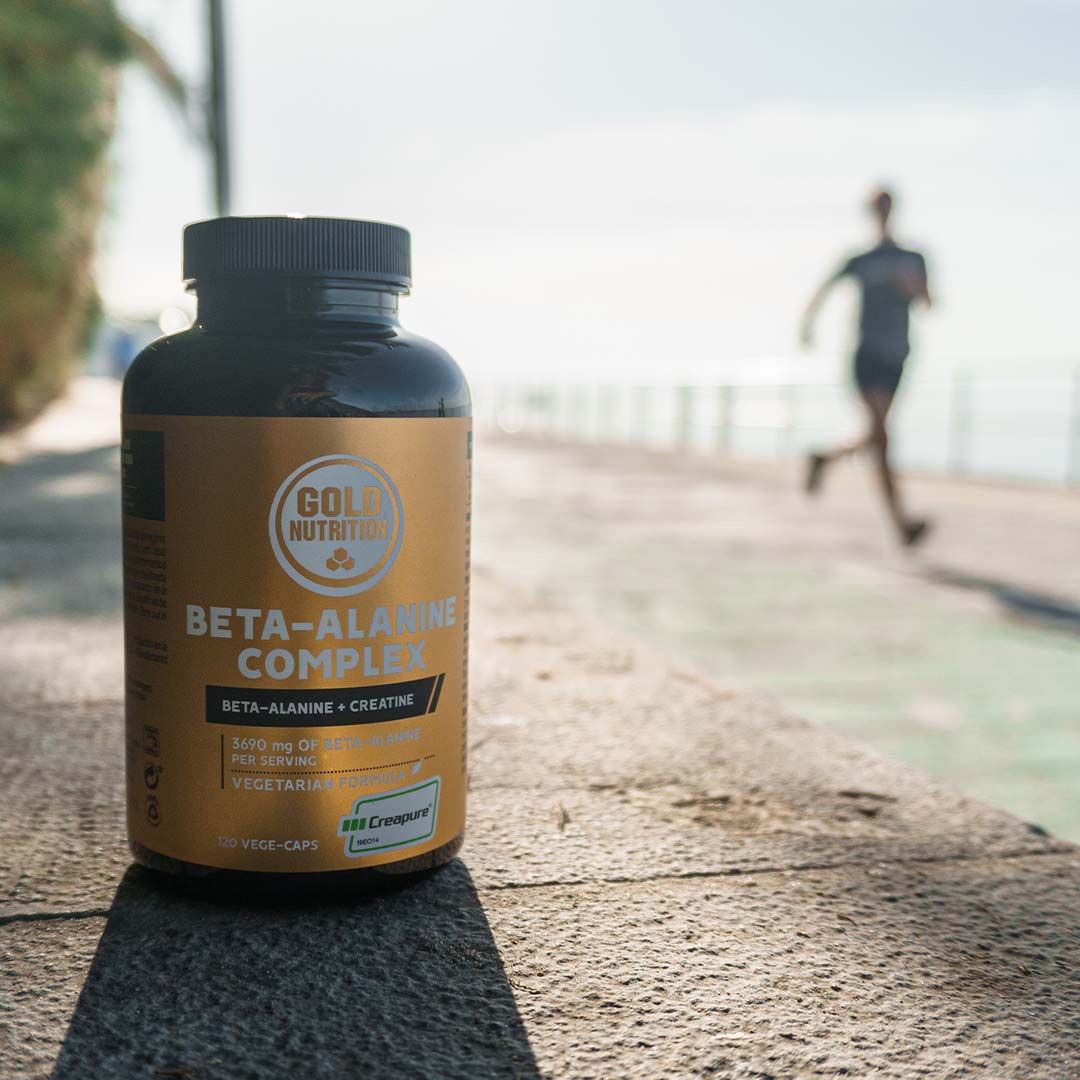 Un frasco de suplemento nutricional etiquetado como Beta-Alanina Complex de Gold Nutrition está en primer plano sobre una superficie de asfalto, mientras una persona corre en el fondo en un camino al aire libre junto al mar | Beta-Alanine Complex