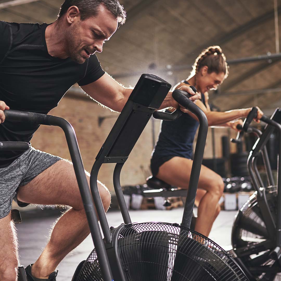 Dos personas haciendo ejercicio intenso en bicicletas de aire dentro de un gimnasio. Están concentradas, trabajando duro mientras pedalean en máquinas diseñadas para entrenamientos cardiovasculares.