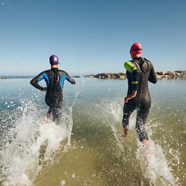 Dos personas corriendo hacia el agua desde la orilla del mar. Están vestidas con trajes de neopreno y gorros de natación, uno con un gorro púrpura y otro con un gorro rojo. Parece que están participando en una competición o entrenamiento de triatlón, ya que el traje es típico para este tipo de actividades. El agua salpica alrededor de ellos mientras avanzan, y el cielo despejado sugiere un día soleado y agradable.