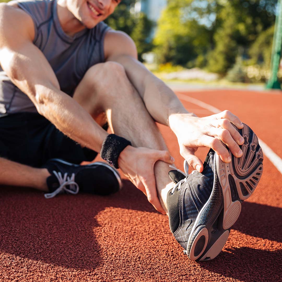 Hombre sentado en la pista de atletismo, sujetando su pierna y tobillo como si tuviera una lesión. Lleva ropa deportiva y una muñequera, con expresión de dolor leve.