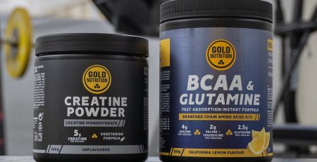 Dois suplementos da GoldNutrition sobre um banco de treino num ginásio. À esquerda, um frasco preto de Creatine Powder (Creatina Monohidratada) e, à direita, um frasco azul de BCAA & Glutamine (Aminoácidos de Cadeia Ramificada com Glutamina). O fundo desfocado mostra equipamentos de treino, incluindo pesos e uma máquina de exercício.
