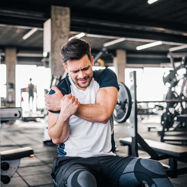 Hombre con dolor en el brazo mientras entrena en el gimnasio. Parece haber sufrido una lesión tras levantar pesas